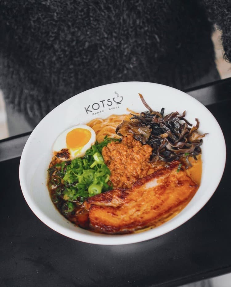 A hand holding a bowl of spicy ramen with the KOTSU® logo on the rim
