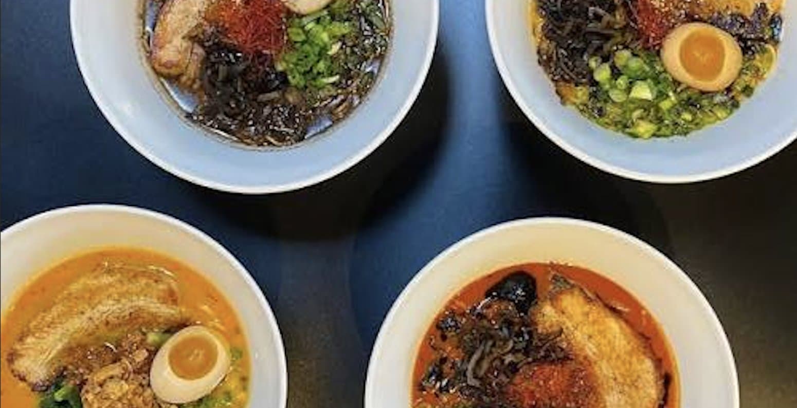 Four white bowls of ramen photographed from above on a dark charcoal surface, with chashu, egg, greens, and chili